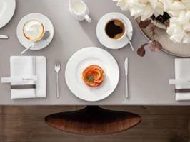 Ein hübsch gedeckter Esstisch mit der Royal-Kollektion von Villeroy & Boch, darunter ein Teller mit Essen, eine Kaffeetasse, ein Cappuccino, Besteck, Servietten und ein Krug Milch. Zusätzlich befindet sich auf der rechten Seite des Tisches ein Blumengesteck.