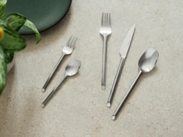 Cinq couverts like. Like. by Villeroy & Boch - fourchette, couteau, cuillère, fourchette à dessert et cuillère à café - sont disposés sur une surface en pierre claire à côté d'une assiette verte et d'une plante.	