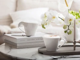 Deux tasses à cappuccino de la collection NewWave sur un élégant plateau. Deux tasses à cappuccino de Villeroy & Boch NewWave, un plateau élégant, des livres décoratifs et une bougie. Les fleurs blanches ajoutent une touche de charme à l'arrière-plan.