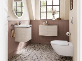 Une salle de bains pour invités avec céramique et meubles de la collection de salle de bains Subway 3.0 de Villeroy & Boch Une salle de bains avec une fenêtre, un sol en galets, un lavabo suspendu et des toilettes. Au-dessus du lavabo se trouve un miroir rond, les murs inférieurs sont recouverts de boiseries et des tableaux encadrés sont accrochés sur les murs supérieurs.