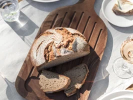Une miche de pain avec deux tranches repose sur une planche en bois Artesano de Villeroy & Boch Une miche de pain avec deux tranches repose sur une planche en bois Artesano de Villeroy & Boch. Sur la table, il y a des assiettes, des serviettes, un verre d'eau, un bol d'olives et un verre de vin.