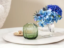 Fleur vases by Villeroy & Boch, a small green and transparent vase with blue flowers, stands on a round white tray with wooden cubes on a glass table.
