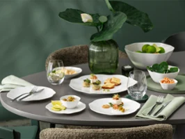 A round dining table set with white tableware from the Pura collection, including starters, glasses, napkins and a centrepiece of green plants.