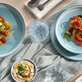 Une table dressée avec des pâtes servies dans des assiettes bleues de la collection Perlemor Aqua. Une table dressée avec des pâtes, de la salade, du houmous, des olives et des couverts sur des assiettes bleues de la collection Perlemor Aqua et une nappe claire.