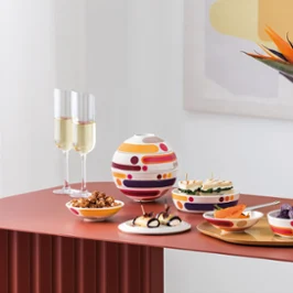 A modern table setting with La Boule Miami, assorted snacks, two champagne glasses, and a white Manufacture vase Collier with orange flowers.