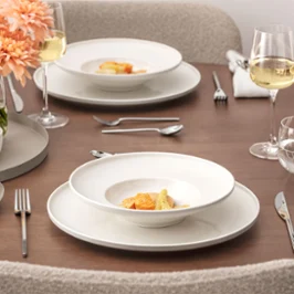 Table à manger dressée de la collection Afina Une table bien dressée de la collection Afina avec deux assiettes garnies de plats gastronomiques, des verres à vin, des verres à eau, des couverts en argent, des serviettes en tissu et un vase avec des fleurs de pêcher sur une surface en bois.