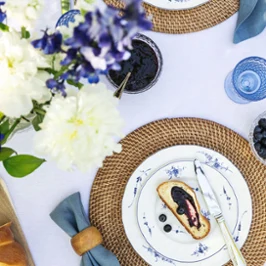 Une assiette Vieux Luxembourg contenant des myrtilles Un dressage de table avec une part de gâteau et des myrtilles sur une assiette Vieux Luxembourg, entourée d’un set de table tressé, de couverts de table et d’une serviette bleue. Sur le côté, on peut voir un vase avec des fleurs blanches et violettes.