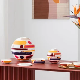 La petite boule miami et la boule miami disposées sur une table rouge Des services de table colorés et composés de plusieurs pièces de la petite boule miami, objets design circulaires de grande et petite taille, sont disposés à titre décoratif à côté d'un vase aux motifs abstraits orné de fleurs orange sur une table rouge.