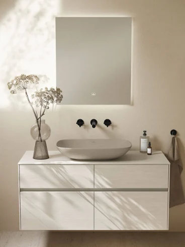 Une salle de bains moderne avec un miroir lumineux, une robinetterie murale, une vasque à poser « Loop & Friends » et un plan de toilette « Collaro ».