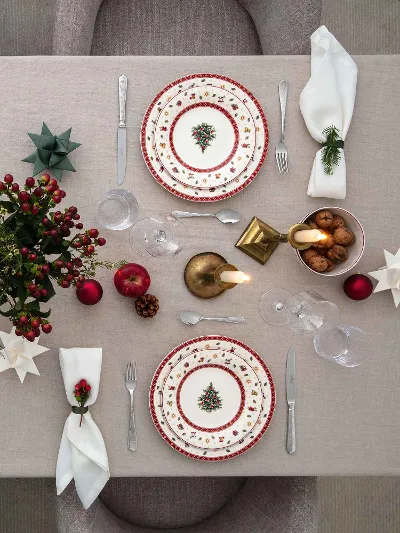 Round Toys Delight plates with Christmas tree patterns in a festive table setting A dining table setting for four people with festive decorations, including Toys Delight plates with Christmas tree patterns, red and gold accents, folded white napkins, candles and a centrepiece with red berries and green plants.