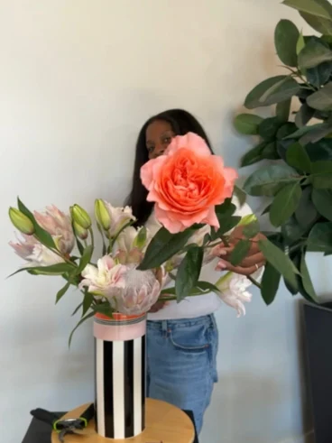 Memphis vase floral arranging Person partially hidden behind a lush coral flower arrangement in a striped vase, with pink lilies and leafy plants creating a warm, serene indoor scene.