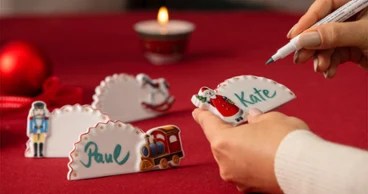 A person is writing ‘Kate’ on a Christmas-themed name card lying on a table, on which there are other name cards and a lit candle.