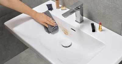 Cleaning a dirty washbasin A hand is cleaning a washbasin in the bathroom with a grey cloth and removing a make-up stain. There are various make-up products on the rim of the washbasin.