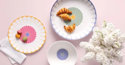 Drei Villeroy & Boch Fleur-Teller mit Croissants und Macarons neben weißen Blumen auf hellrosa Untergrund.