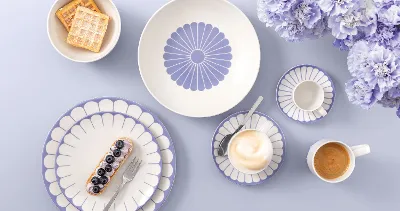 Tischgedeck mit Villeroy & Boch Fleur Bleu-Tellern, Kaffee, Dessert, Waffeln und hellvioletten Blumen auf hellem Untergrund.

