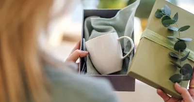 Woman holding a gift box with a Crafted ceramic mug and a green cloth decorated with a eucalyptus branch.