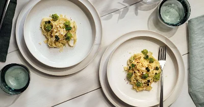 Deux assiettes Crafted avec du brocoli et de la sauce sur une table blanche, accompagnées de verres à eau et d’une petite plante en pot.