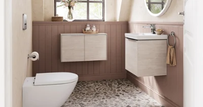 Une salle de bains avec une fenêtre, un sol en galets, un lavabo mural et des toilettes. Au-dessus du lavabo se trouve un miroir rond, les murs inférieurs sont recouverts de boiseries et des tableaux encadrés sont accrochés sur les murs supérieurs.