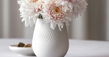 Vase Manufacture blanc avec des chrysanthèmes rose pâle sur une table, à côté d'une petite coupe avec des pierres.