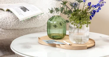 Une table basse ronde avec un vase Villeroy & Boch Fleur contenant des fleurs bleues et vertes. En arrière-plan, un magazine ouvert est posé sur une chaise blanche et moelleuse.