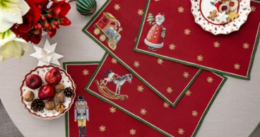 Toy's Delight Gobelin placemats with Christmas motifs Overhead view of four red Toy's Delight tapestry placemats featuring a nutcracker, rocking horse, train, and Santa Claus on a dining table, decorated with Christmas cookies, apples, walnuts, and festive ornaments