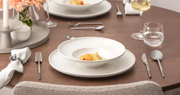 Gedeckter Esstisch der Afina Kollektion Ein ordentlich gedeckter Esstisch der Afina Kollektion mit zwei Tellern mit Gourmetgerichten, Weingläsern, Wassergläsern, Silberbesteck, Stoffservietten und einer Vase mit Pfirsichblüten auf einer Holzfläche.