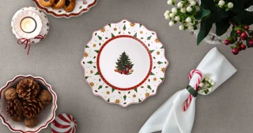 A festively set table with a decorated plate featuring a Christmas tree motif, cookies, fruit, candles, napkins, and a flower arrangement. 