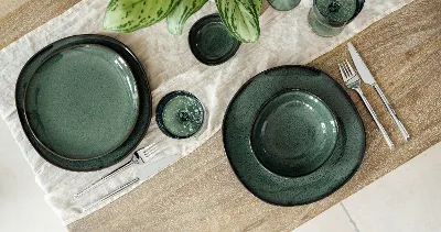 Table en bois élégamment dressée avec de la vaisselle en céramique "Lave Vert", des couverts en argent "like" de like. by Villeroy & Boch et des verres Like assortis sur une serviette en tissu crème. Une plante verte avec des accents jaunes complète l'arrangement raffiné.