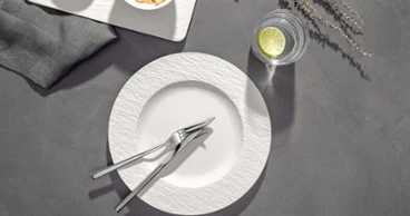 Assiette blanche vide avec fourchettes et couteaux croisés, un verre d'eau avec du citron vert et une serviette de table sur une surface grise.
