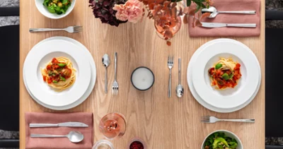 Table de salle à manger avec deux assiettes royales de pâtes, deux salades, des couverts, une bougie et une boisson. Table de salle à manger avec deux assiettes royales de pâtes, deux salades, des couverts, une bougie et une boisson. Les assiettes Villeroy & Boch sont posées sur des serviettes roses et une composition florale rose est disposée sur la table.