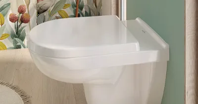 Modernes Badezimmer mit wandmontiertem O.novo-WC, hellgrünen Wänden und geblümter Tapete. Neben dem WC befindet sich ein kleiner runder Tisch mit gefalteten Handtüchern.