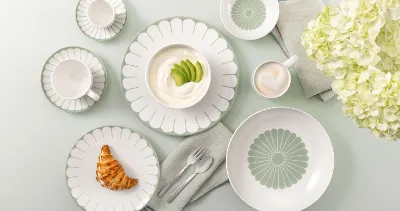 Tischgedeck mit Villeroy & Boch Fleur Vert-Tellern, Croissant, Joghurt, Kaffee und weißen Blumen auf hellgrünem Hintergrund.