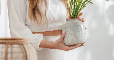 Une femme vêtue de blanc remet un vase en céramique Lave avec des fleurs vertes et jaunes, qui est posé sur une table en bois.