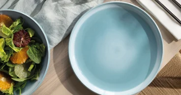 Une assiette bleue nacrée Aqua vide est posée sur une table en bois, à côté d'un saladier, de couverts et d'une serviette.