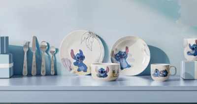 Una serie di piatti e tazze della collezione Disney Stitch sono decorati con motivi dei cartoni animati ed esposti su uno scaffale azzurro insieme ad altri utensili.