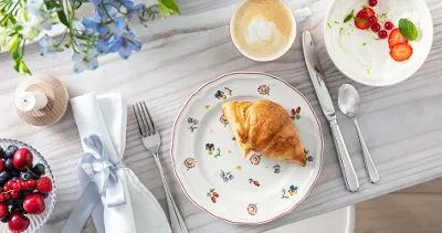 A croissant on a flower plate with coffee, berries, yogurt, cutlery and a napkin with a bow.