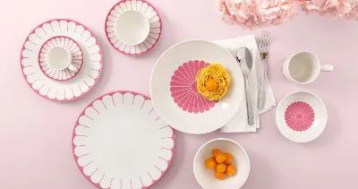 Tischgedeck mit Villeroy & Boch Fleur Cassis-Tellern, Nudeln, Clementinen und Blumen auf rosa Hintergrund.