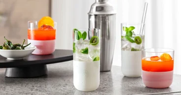 Quatre cocktails colorés dans des verres Manufacture blancs sur une surface en marbre, avec un shaker à cocktail en arrière-plan et un petit bol de paprika sur un plateau.