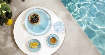 Un vassoio rotondo con stoviglie della collezione Perlemor Aqua Un vassoio rotondo con stoviglie della collezione Perlemor Aqua e un bicchiere d'acqua, sul bordo di una piscina.