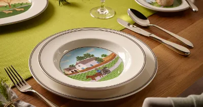 A table setting with a bowl depicting a farm scene, surrounded by cutlery, a napkin and a bread plate.