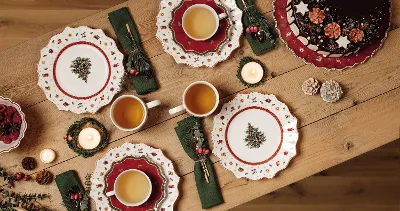 A wooden table with Christmas plates, green napkins, cups of tea, candles and a chocolate cake.