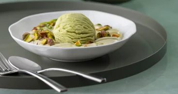 Une coupe à dessert Pura contenant de la glace à la pistache, des pistaches hachées et de la crème, posée sur une assiette blanche avec des couverts à côté.