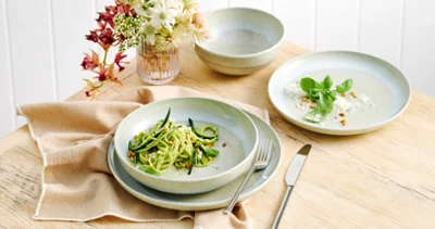 aus-perlemoralga-indoor-food-nopeople-dinnerset7069 The Perlemor Alga dinner set for one, featuring a plate filled with fresh zucchini pasta, rests on a brown linen tablecloth atop a rustic wooden table, accompanied with silver cutlery.