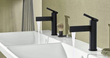 Une salle de bains moderne avec deux robinets noirs au-dessus d’un lavabo blanc. Un blaireau et une serviette sont posés sur la tablette. 
