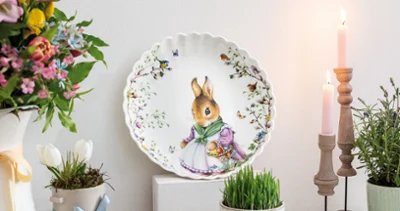 The Spring Fantasy decorative plate by Villeroy & Boch with a bunny motif. The Spring Fantasy decorative plate by Villeroy & Boch with a bunny motif is displayed on a shelf, surrounded by flowers, candles, and a wooden egg.
