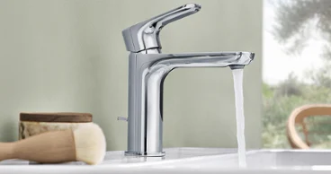 Sur un lavabo blanc O.novo est installée une robinetterie moderne en chrome avec de l’eau qui coule. À côté, se trouvent une brosse et un récipient en bois.