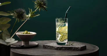 Un verre La Divina avec des tranches de concombre et du romarin, sur une surface en pierre avec un fond vert. Une paille en verre Artesano est plantée dans la boisson et une coupe d’olives est posée à côté.
