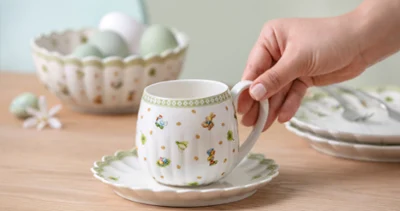 A hand holds a Easter Delight teacup on a saucer, with a bowl of eggs and plates in the background on a wooden table.