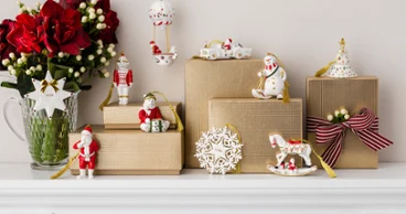 Christmas Classics Ornament Collection 2026 Several wrapped presents, Christmas decorations and a vase of red flowers are arranged on a white mantelpiece.