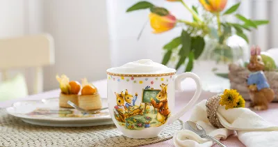 The decorative mug with foam crown from the Villeroy & Boch Annual Easter Edition, decorated with a bunny motif, stands on a table next to a dessert plate, a flowered tablecloth and a bunny figurine. Tulips bloom in the background.